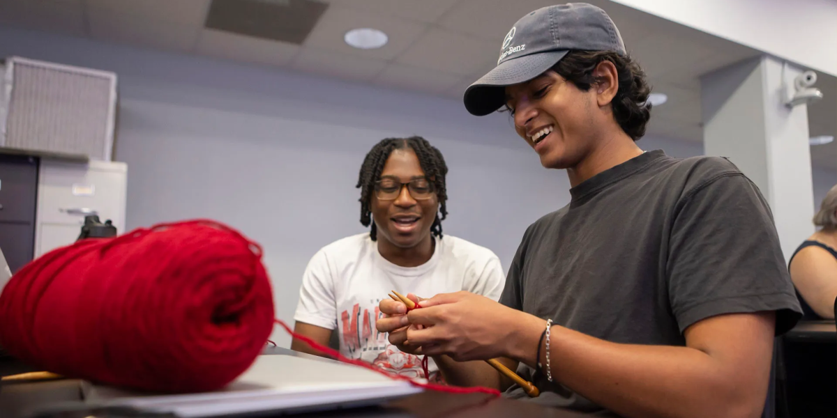 Two male students are seated at a table in a classroom. The student in the foreground, wearing a dark baseball cap, is smiling while knitting with red yarn. The student seated behind him is also smiling and watching.
