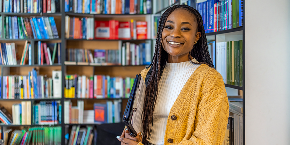 Smiling woman holding a book stands between tall library shelves filled with books, suggesting an academic or learning environment.